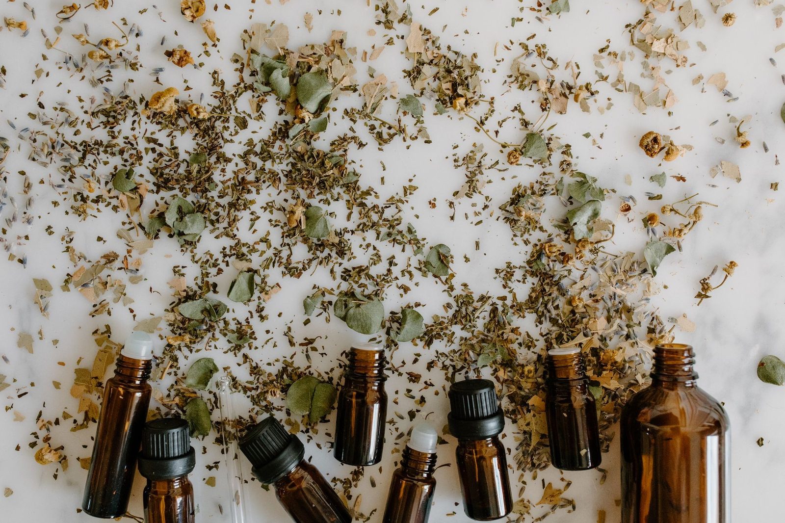 Frascos de vidrio con aceites esenciales rodeados de hierbas secas sobre fondo blanco